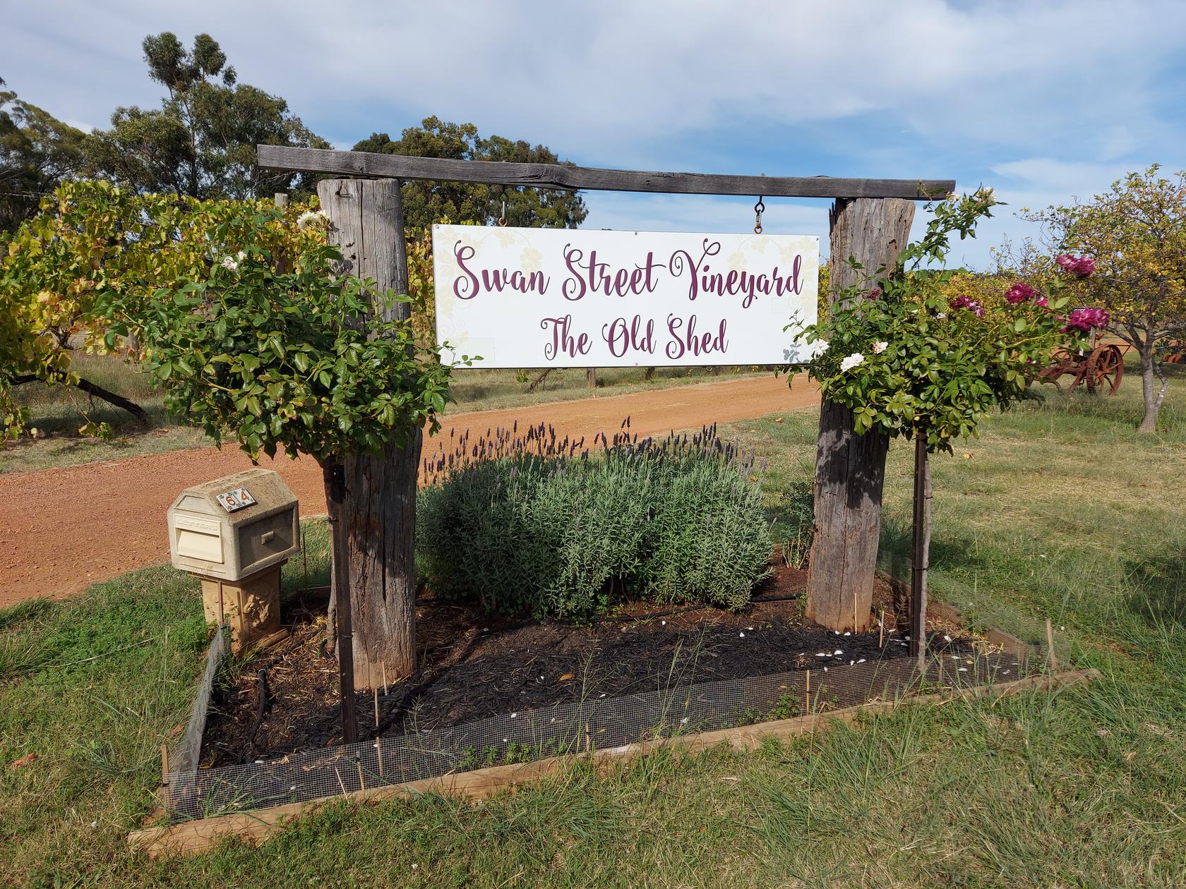 The Swan Street Vineyard entrance sign hung between two timber posts at the property driveway, surrounded by roses and lavender.