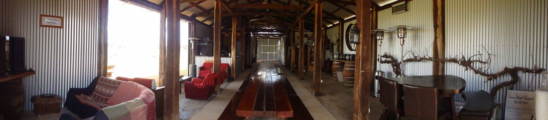Wide panorama inside the shed showing the timber-post corridor, the long jarrah table, lounge seating and warm interior light.