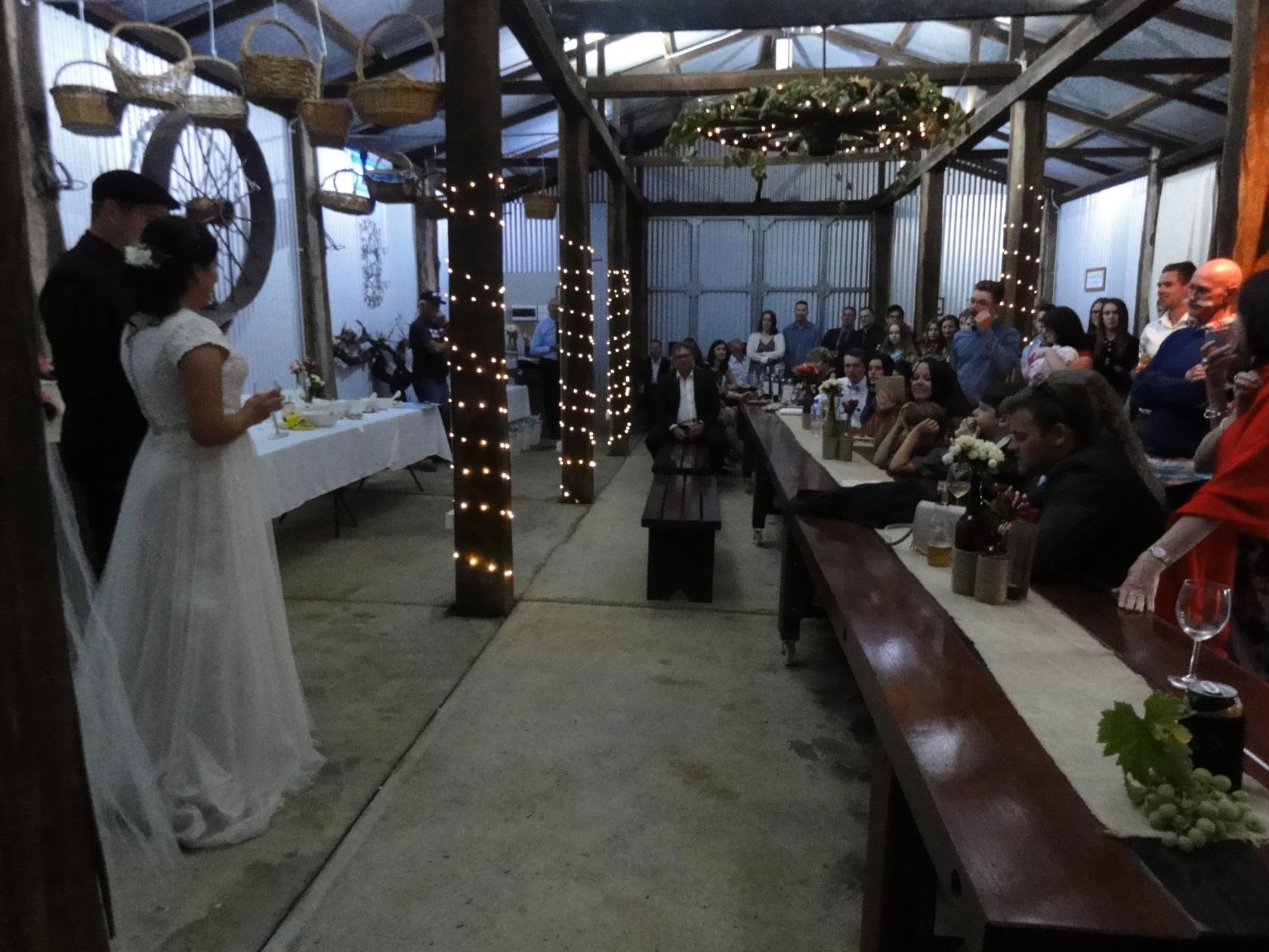 A wedding ceremony inside the shed at dusk, the couple standing before guests seated along the long jarrah table strung with fairy lights.
